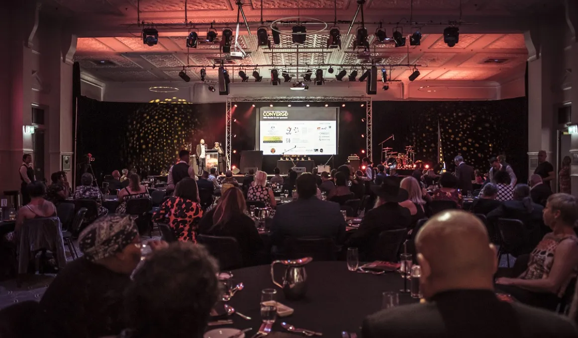 Large group of Aboriginal and Torres Strait Islander people sitting at tables in a darkened room. In the background is a stage and on the ceiling, stage lights.