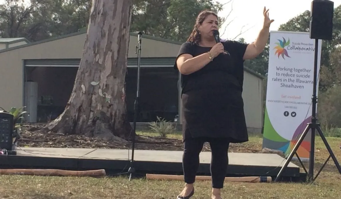 Aboriginal woman in black clothing holds a microphone as she stands on grass. In the background is a large tree, a shed, a sign, a speaker on a stand and some didgeridoos laying on the grass.