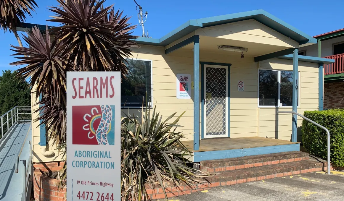 A yellow building with blue trim and a portico has a door at centre with windows either side. In front is a hedge at right and at left, some trees and a bush. In front of the trees is a large sign with the words: SEARMS Aboriginal Corporation.