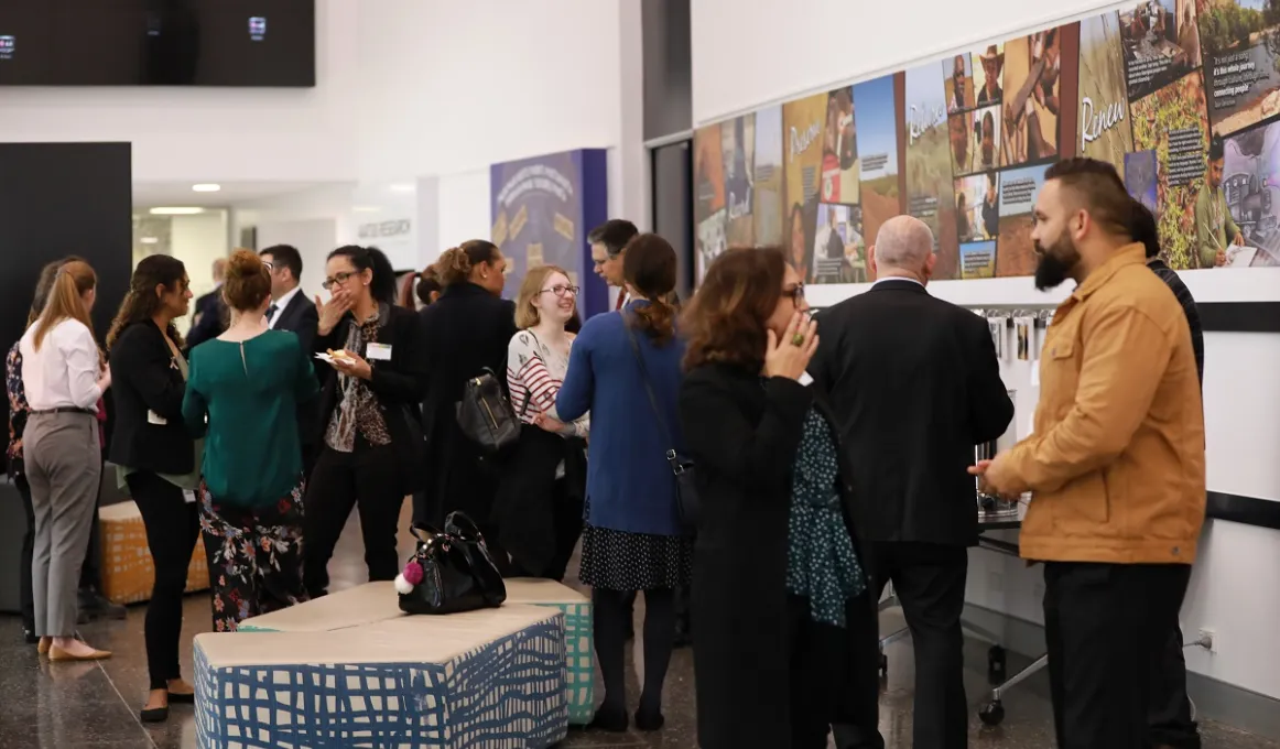 Group of Indigenous people, young and old, talk amongst themselves in a room with art on the walls, a white ceiling and dark tiled floor.