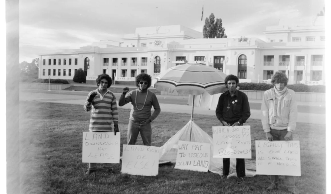 Four men in casual wear stand on grass and either side of a beach umbrella. In front of them are small hand written signs. In the background is a road and then a white building with many windows.