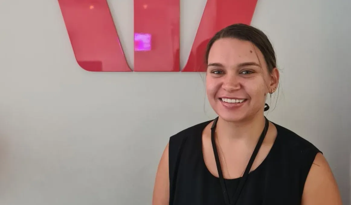 A woman in black dress with hair pulled back stands in front of a red sign in the shape of a W.