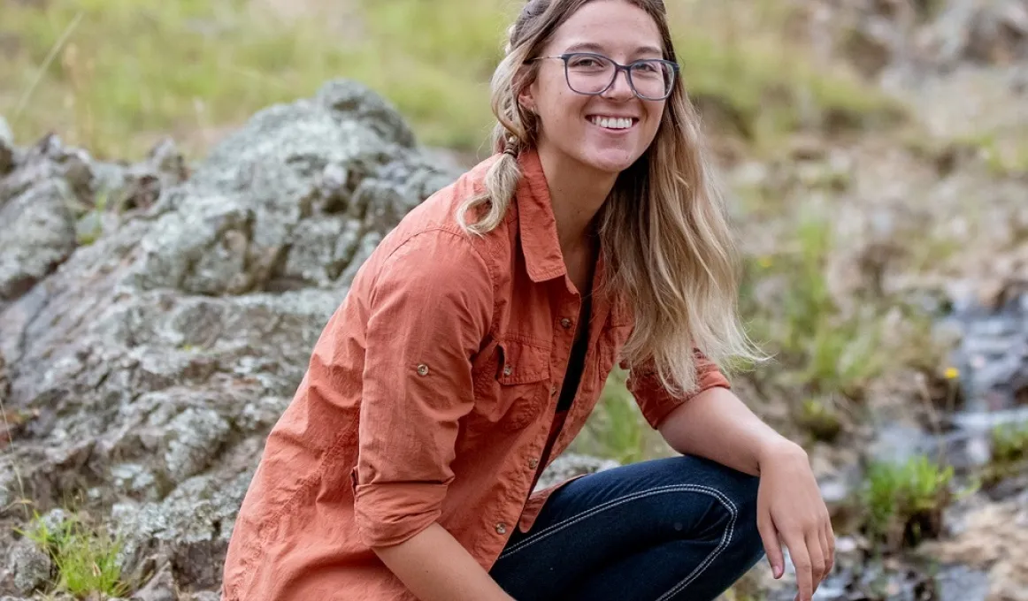 A woman with long blonde hair dressed in an ochre coloured shirt and blue jeans squats on some rocks and looks to camera. In the background are rocks and grass.