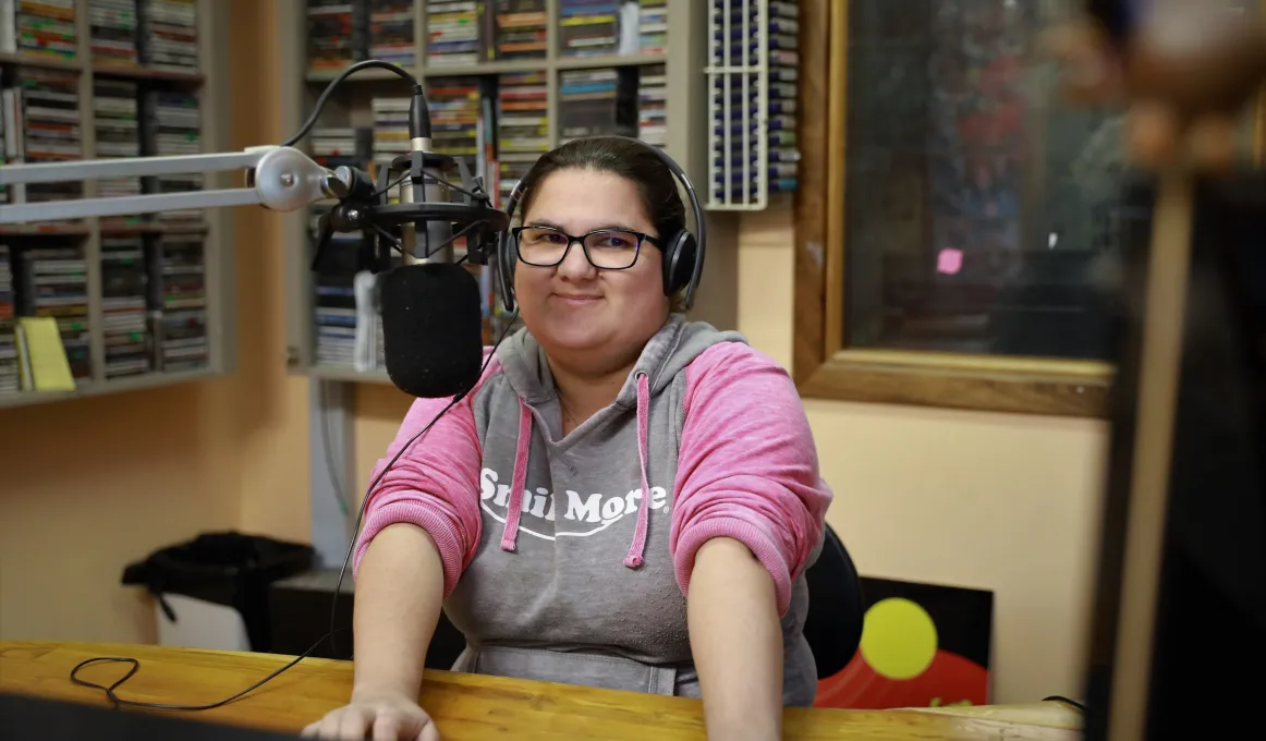 Aboriginal woman wearing glasses and a grey and pink top sits at a desk in front of a microphone. In the background is a rack of discs, a window and an Aboriginal flag.