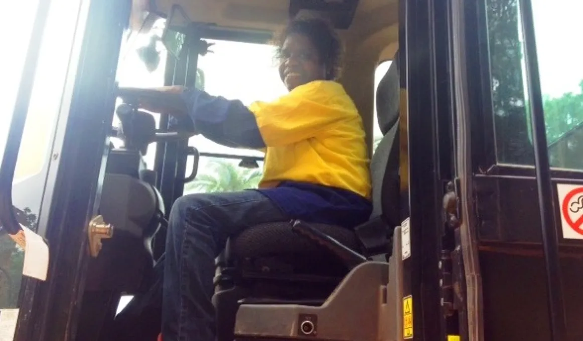 Indigenous woman - Sandra Gillman - sits in the cab of a front end loader wearing yellow and blue safety clothing.