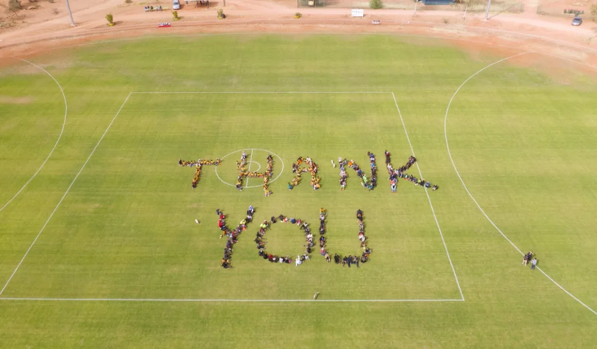 A bird’s eye view of people on a green oval. They stand close together and form the words ‘Thank you’. In the background is a brown road, buildings and more people.