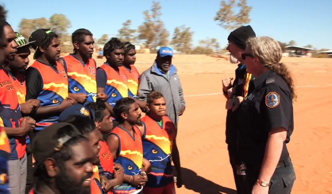 Group of Aboriginal footballers and others associated with the team on an ochre coloured football field.