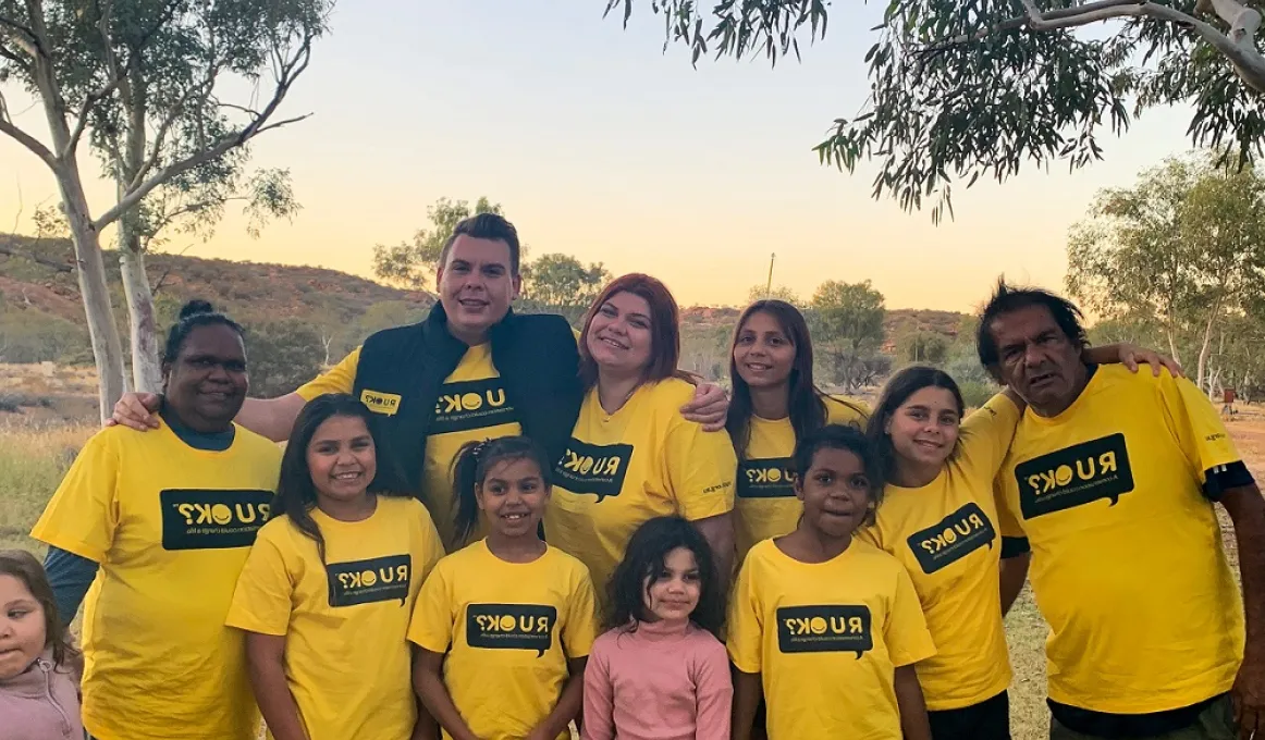 Group of Indigenous adults, youth and children in yellow t-shirts with the letters RUOK on them. Two young girls are dressed in pink tops. In the background is a small hill and trees.