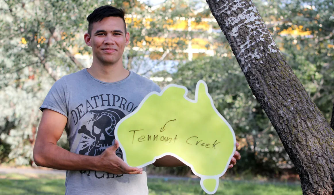 Young Indigenous man wearing grey t-shirt standing beneath a tree and holding a cardboard cut-out of Australia featuring a dot with an arrow to the words Tennant Creek.