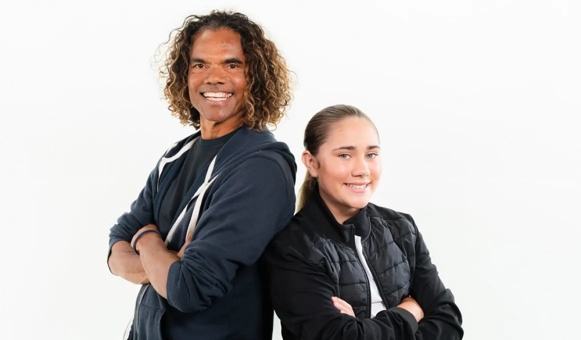 A smiling man in dark top with long, wavy hair stands back to back with a young woman in a dark top and hair pulled back.