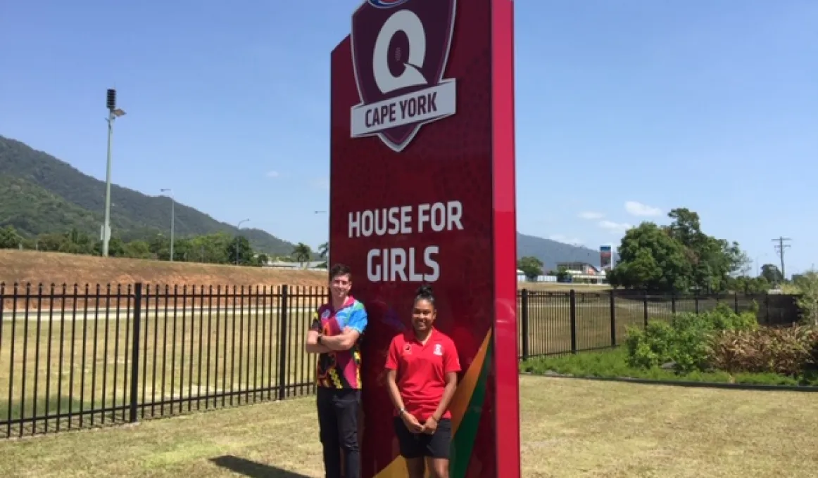 Adult male in multi-coloured shirt and adult female in red shirt stand in front of a large red sign which says AFL Cape York House for Girls. In the background is grass, a road, some buildings and hills.