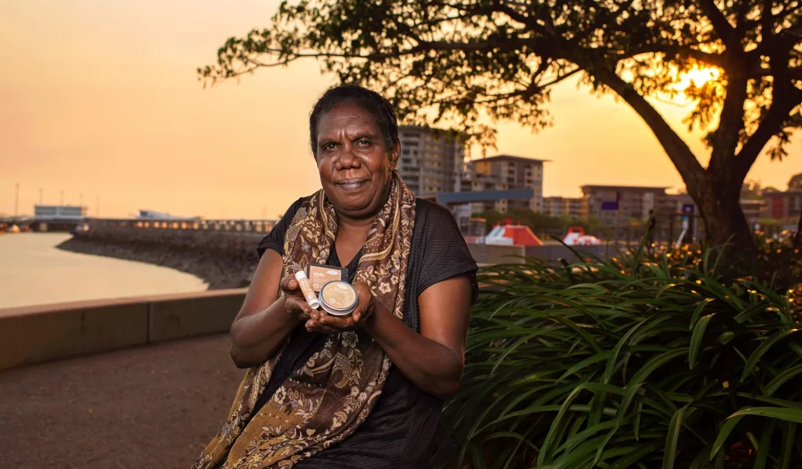 An Aboriginal woman in dark dress and patterned scarf sits next to a bush and holds three small containers in her hands. In the background is water, a wharf, a tree and buildings.