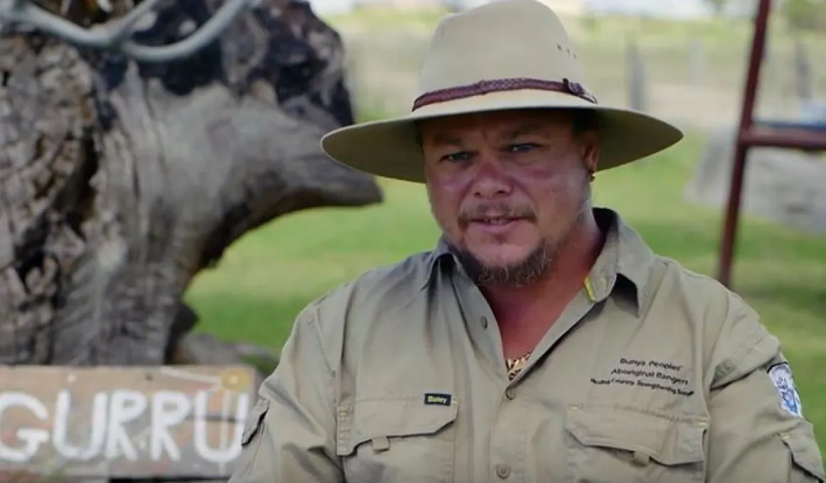 An Aboriginal man wears a broad brimmed hat and a khaki uniform. In the background is a tree, grass and metal structure.