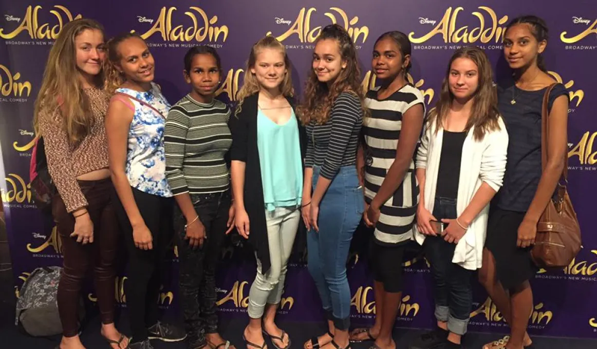 Eight young Indigenous women standing on a carpeted floor in front of a poster wall with multiple displays of the word ‘Aladdin’.