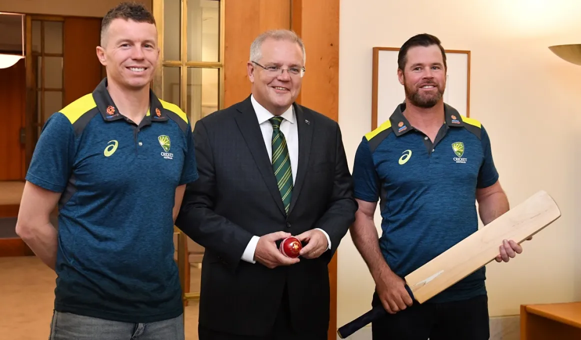 Three men stand in a room. In the background is a white wall and a doorway through to neighbouring room. The man on the left wears a blue polo shirt and blue jeans. The man in the middle wears a dark suit. The man on the right wears darks trousers.