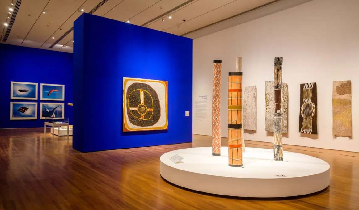 A gallery with a beige ceiling and shiny board floor features two blue walls. In the gallery are different pieces of Indigenous art.