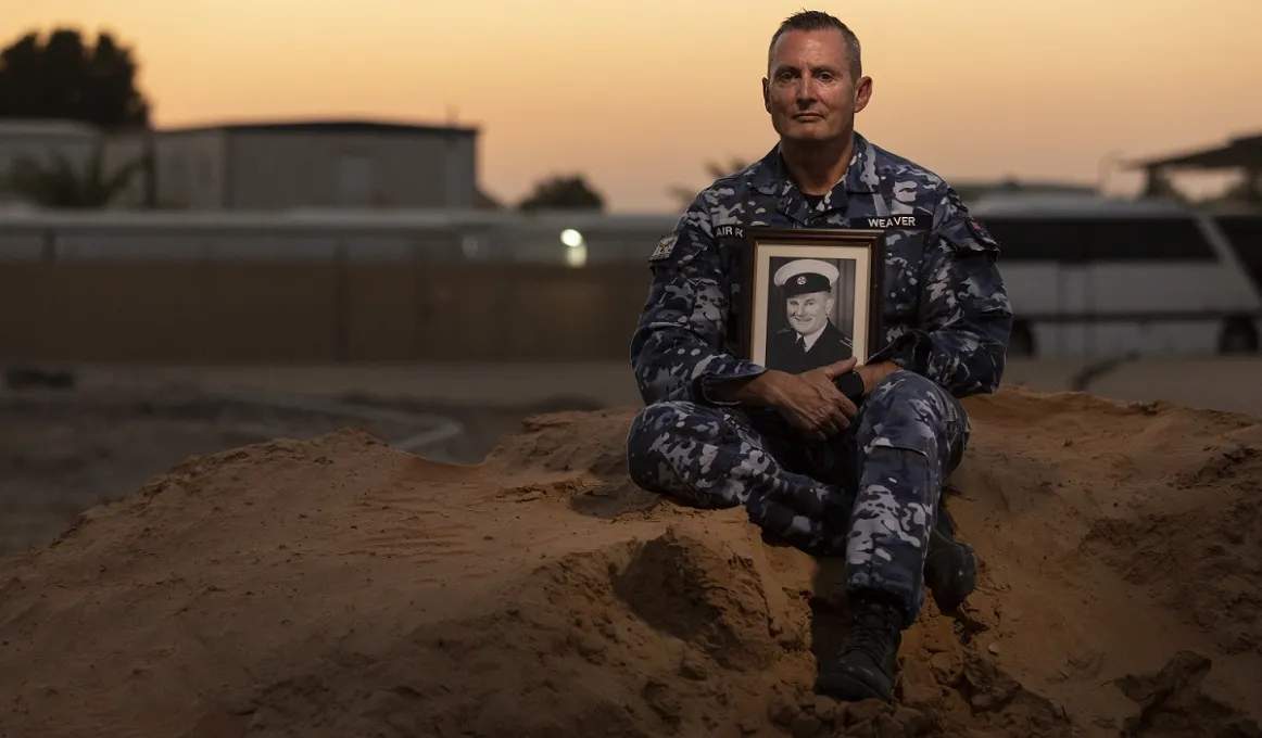 A man dressed in an air force uniform and holding a photo of another man in uniform sits on a dirt mound with a setting sun and buildings in the background.