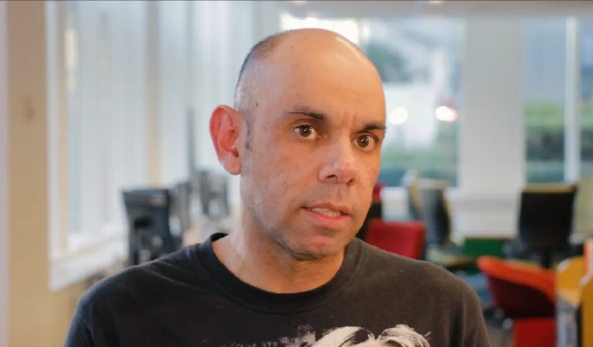 Balding man in black t-shirt in foreground with office furniture and windows in the background.