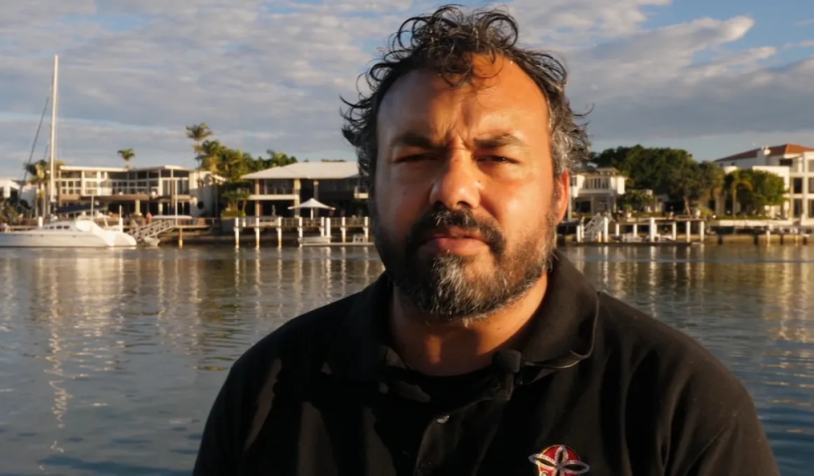 Indigenous man in black polo shirt faces camera. In the background is water and behind that, a boat, wharfs, buildings, trees and a cloudy sky.