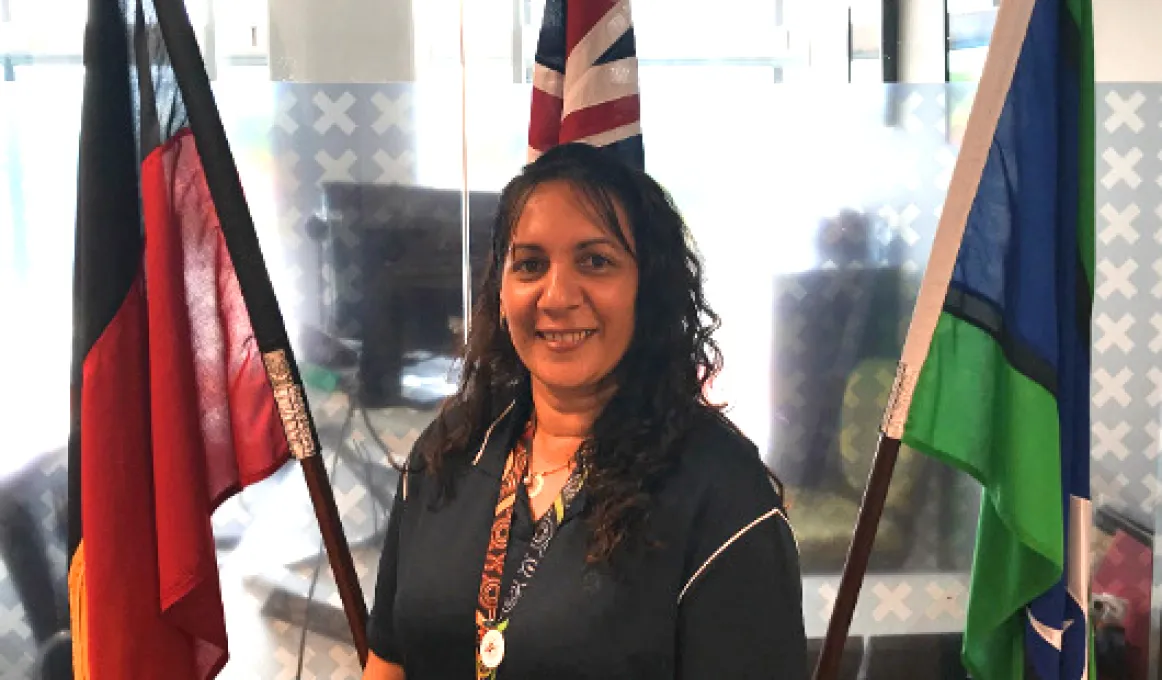 Indigenous woman with long black hair wearing black clothing stands in front of the Aboriginal, Australian and Torres Strait Islander flags. In the background is a glass wall behind which is an office.