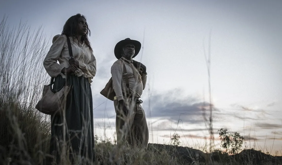 Indigenous woman in white shirt and dark skirt with handbag over shoulder stands next to Indigenous man with hat, light shirt and pants standing in the outback with sunset behind.