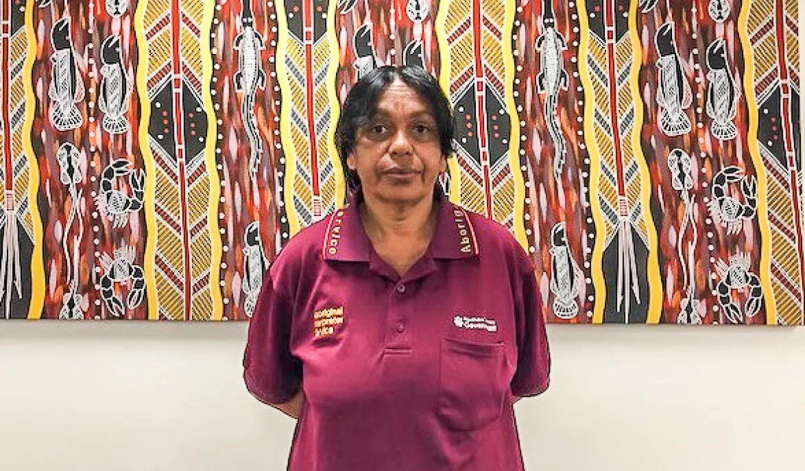 Aboriginal woman in maroon polo shirt stands in front of an Aboriginal painting of featuring vertical patterns of lines and animals and spears and other figures.