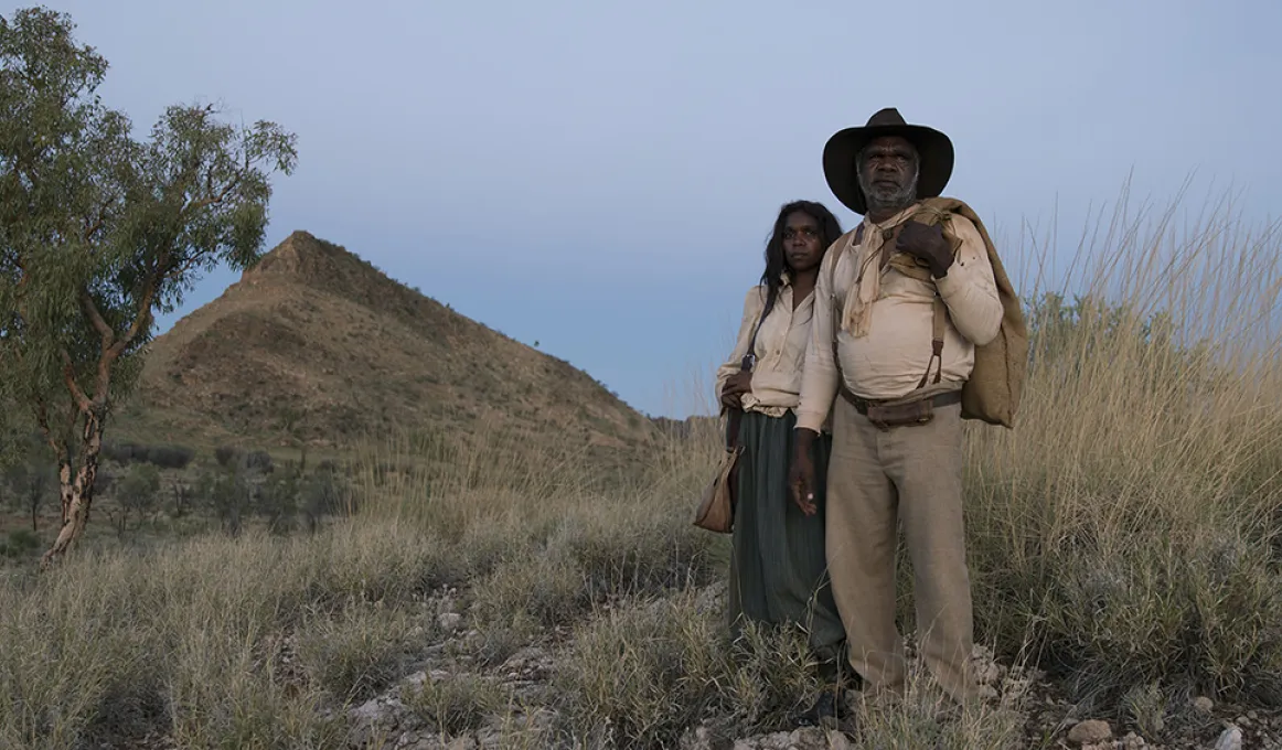 Aboriginal man and woman dressed in 1800s clothing stand on a rock and grassy hill. In the background are trees, a mountain peak and blue sky.