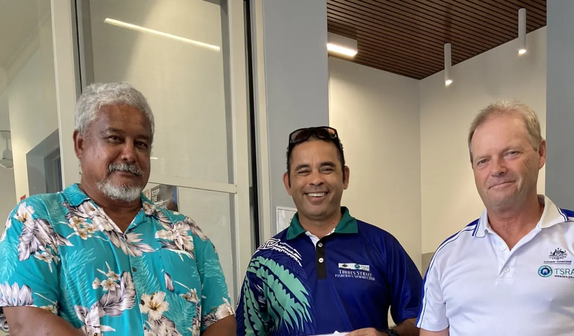 Three men in casual shirts stand shoulder to shoulder and smile at the camera. In the background is a ceiling, walls and windows.