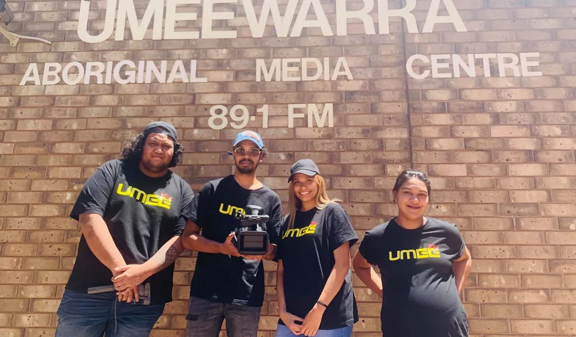 Four Aboriginal young adults (2 men and 2 women) in black t-shirts with UMEEtv printed on them stand in front of a brick wall. On the wall are the words: Umeewarra Aboriginal Media Centre 89.1 FM