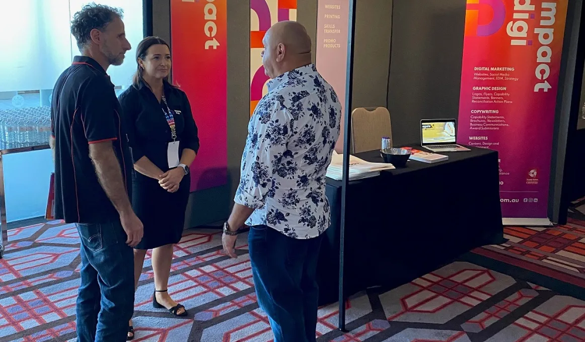 Two men and a woman stand in a circle facing each other on multi-coloured carpet in a room. Behind them are banners with the text Impact Digi, a chair and a table on which is a laptop and some brochures.