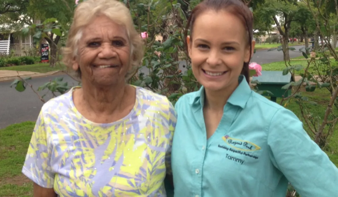 Elderly Indigenous woman in yellow and grey dress stands with arm around young Indigenous woman dressed in aqua coloured shirt in front of a flowering bush in a suburban street.