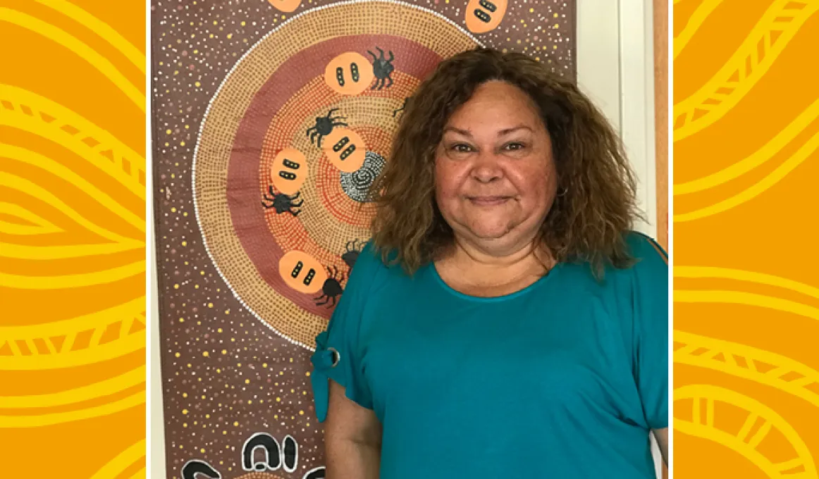 Indigenous Primary School teacher standing front of Indigenous artwork of Honey ants and dots in circles on a brown background.