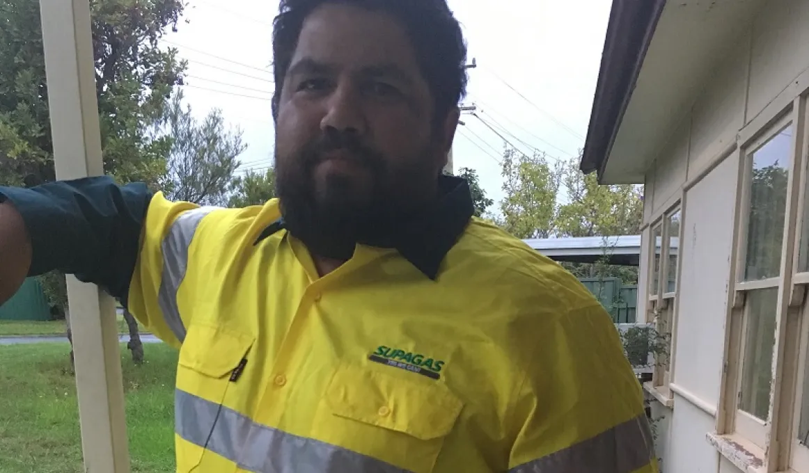 An Aboriginal man with beard and wearing a yellow safety shirt stands next to a beige coloured building. In the background is green grass, trees and another building.