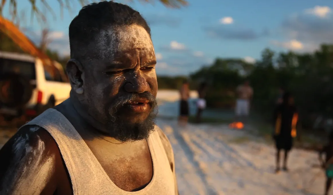 Aboriginal man Timmy Burarrwanga in foreground with others in background outdoors, standing on sandy soil.