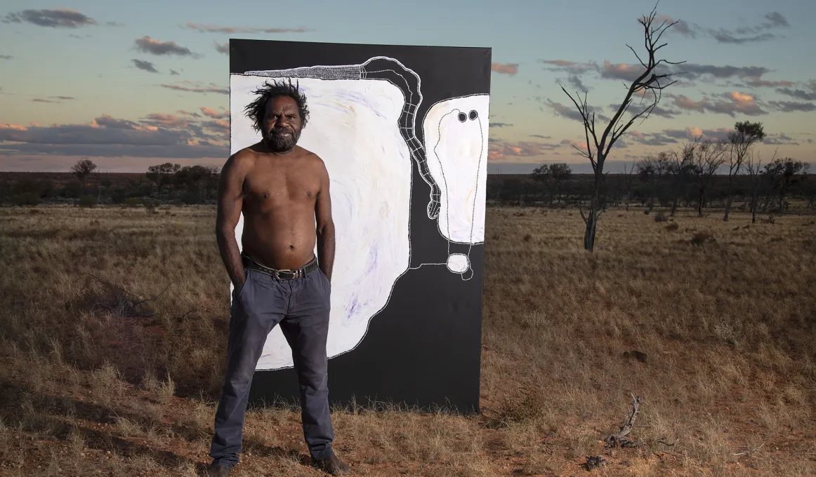 A man wearing dark pants and without a top stands on semi-arid and grassy soil. Behind him is a large black and white painting. In the background are a few trees, a flat landscape and darkening sky.