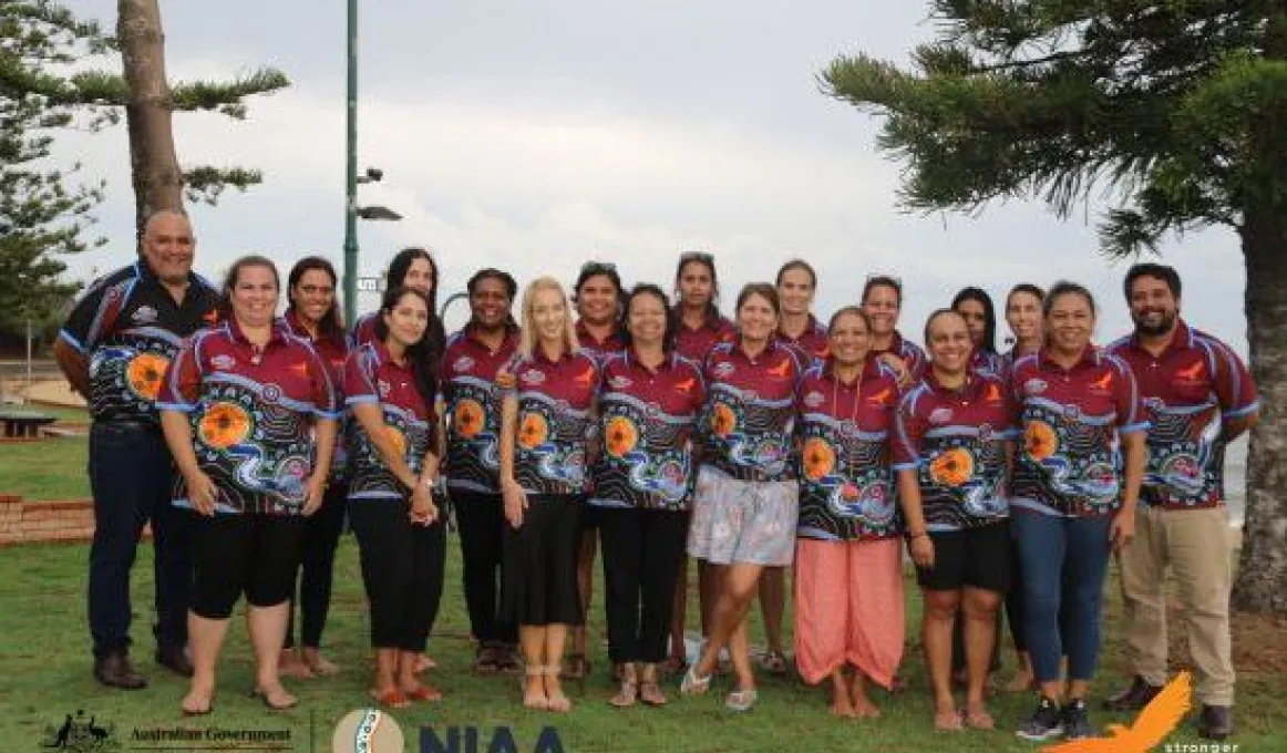 Large group of Indigenous adults wearing the same polo shirt with Indigenous designs stands on grass. In the background are trees, a small wall and cloudy sky.