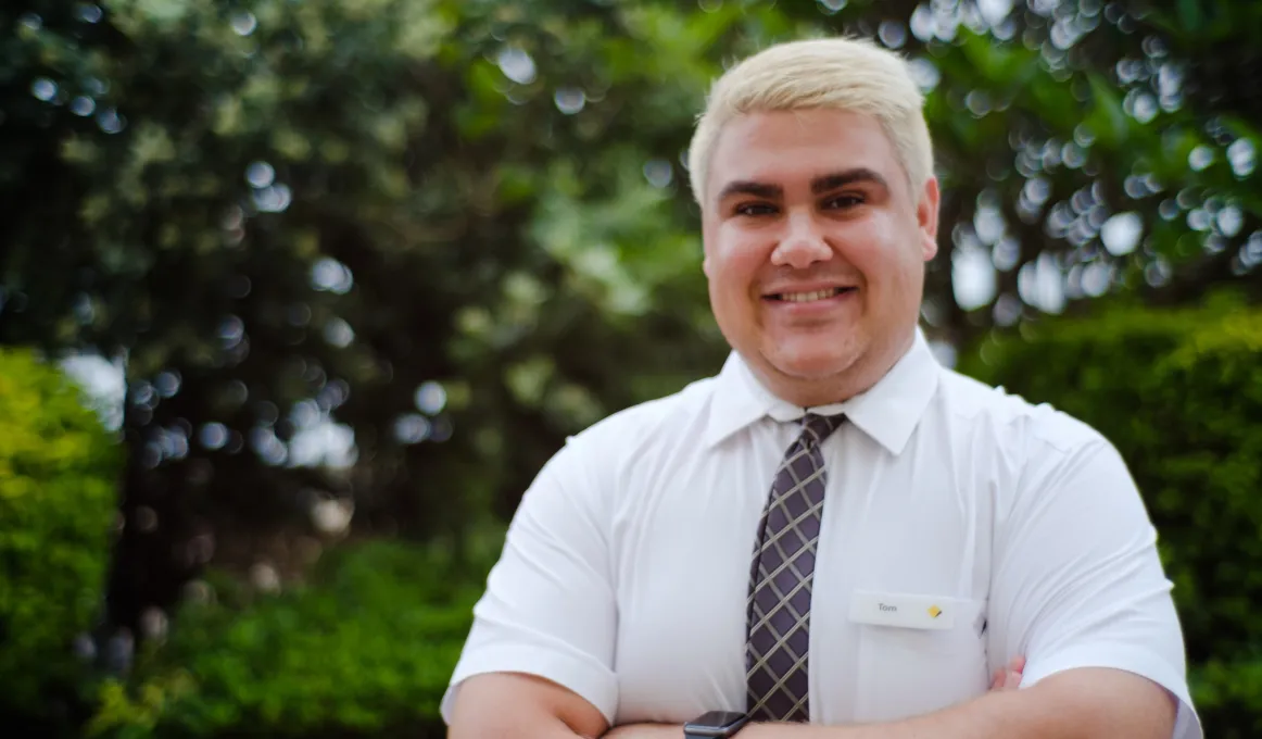 Young adult male in white shirt and tie with short blonde hair stands with arms folded in front of trees and bushes.