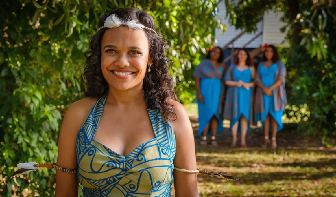 A bride is in traditional Tiwi wedding dress, of bright blue and lime green and is smiling towards the camera. She is outside on a sunny day and there are 3 bridesmaids in blue dresses in the background.