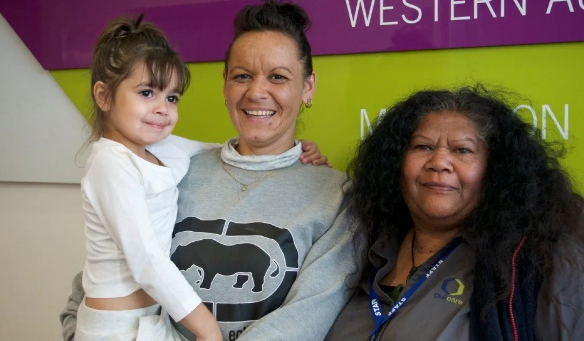 Two Indigenous adult women, one dressed in light grey shirt and holding a child dressed in white clothes, the other in a dark grey shirt.