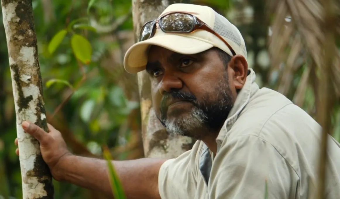 Middle aged Indigenous man with beard wearing a pale shirt and hat with sunglasses on his head leans against a small tree. In the background is foliage.