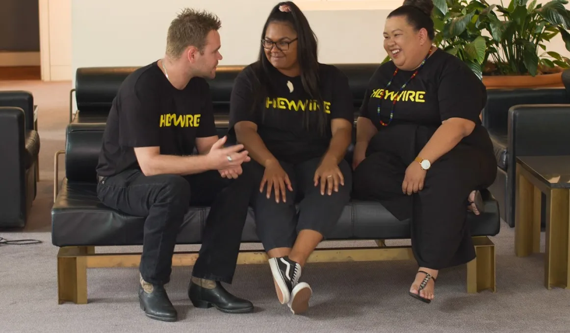 One Indigenous man and two Indigenous women dressed mainly in black, sit on a black couch. In the background is a white wall and some indoor plants. On the floor is a grey carpet.