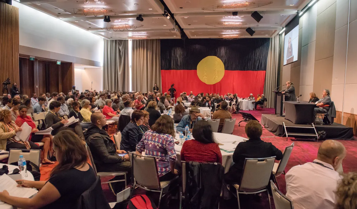 Large gathering of people in a hall, one standing at a rostrum and most seated at tables or on seat rows. The carpet is red, the walls are beige and the curtains are brown. Covering the far end wall is the Aboriginal flag.