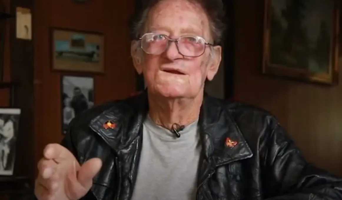 A mature man in leather jacket and wearing glasses sits in a room. In the background is a wall covered in photos and other images.