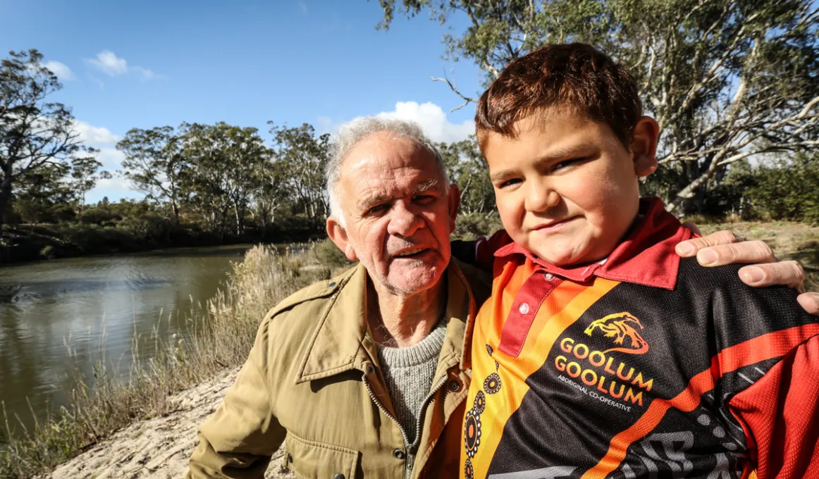 An Aboriginal man in brown jacket and boy in red, black and yellow shirt, stand near a river behind them. Also in the background are trees, grass and a blue sky.