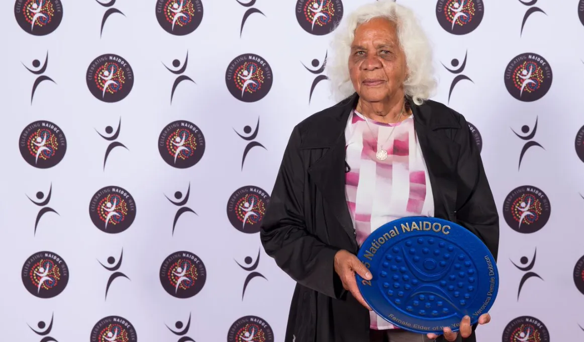 Elderly Indigenous woman, dressed in dark brown over coat and pink and white blouse, holding a blue award in the shape of a plate.