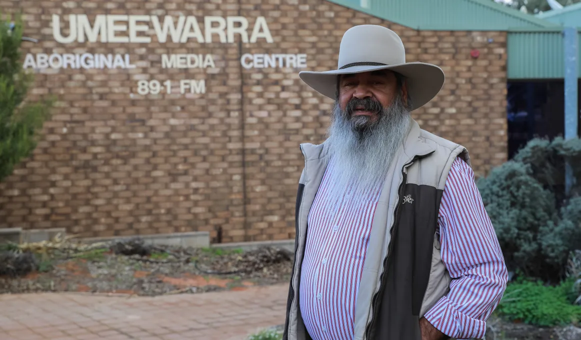 Aboriginal man with long white beard wearing a hat, striped shirt and vest stands in front of a brick building with the following words on the wall: Umeewarra Aboriginal Media Centre 89.1 FM.