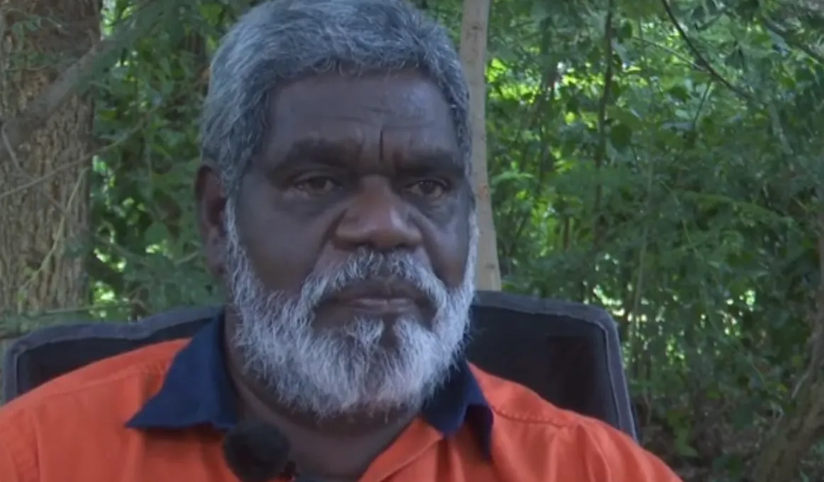 Man in orange shirt with dark blue collar, sits in black chair in front of trees.