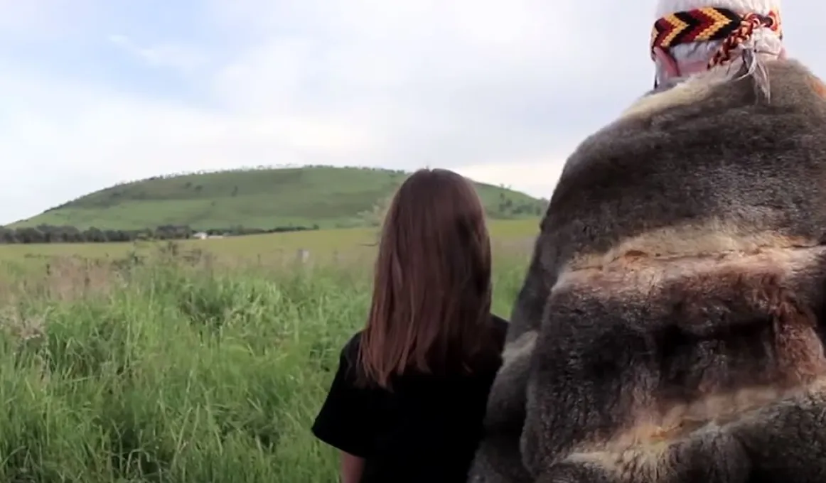 In the foreground we see the backs of an elderly man in a fur cloak and a young girl in a black shirt. Immediately in front of them is long grass and in the background is a large grass covered hill.