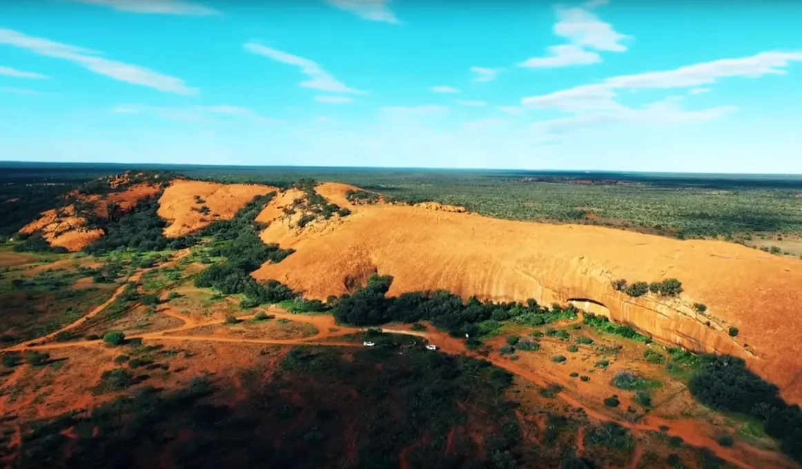 Large ochre coloured rock lies on a plain of ochre coloured soil and bushland.