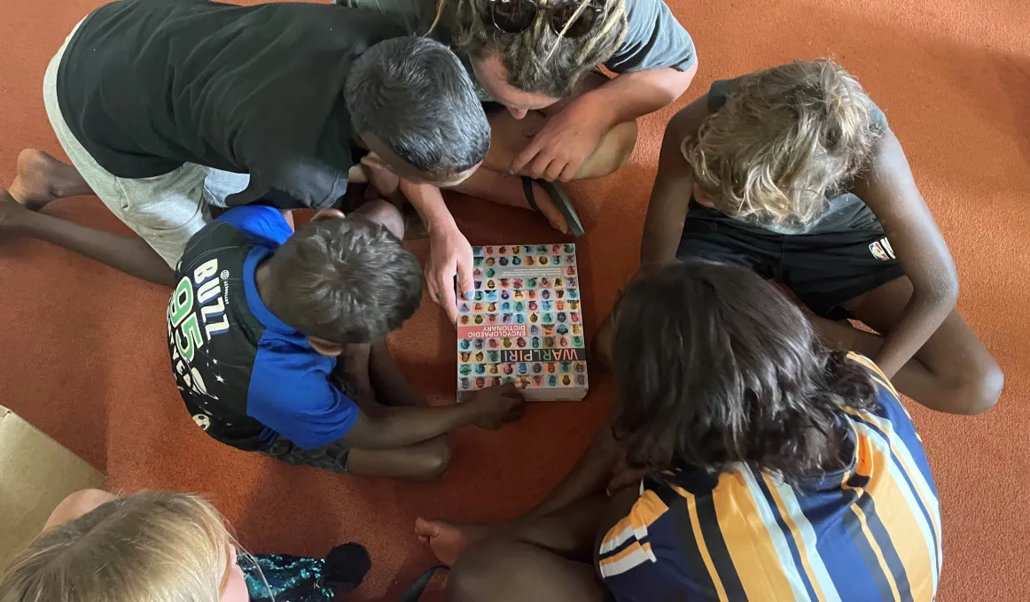 The arrival of the Warlpiri Encyclopaedic Dictionary in Yuendumu, Northern Territory. Five children and an adult are bent over the Warlpiri Encyclopaedic Dictionary. The cover of the dictionary has photos of people who have contributed to it.